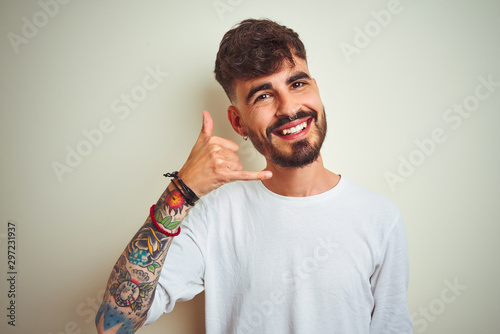 Young man with tattoo wearing t-shirt standing over isolated white background smiling doing phone gesture with hand and fingers like talking on the telephone. Communicating concepts.