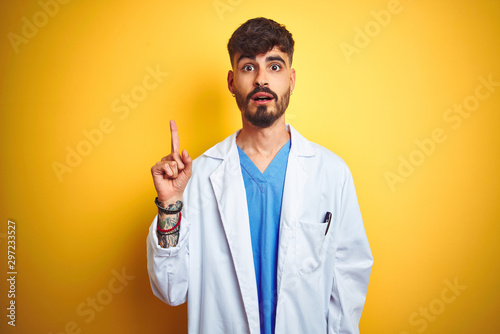 Young doctor man with tattoo standing over isolated yellow background pointing finger up with successful idea. Exited and happy. Number one.