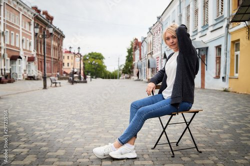 Blonde funny pretty girl sitting on chair in Central part of old city. Walk in downtown. Portrait of girl on street