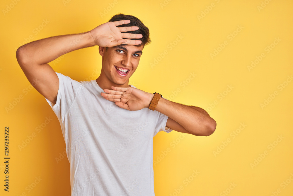 Young indian man wearing white t-shirt standing over isolated yellow background Smiling cheerful playing peek a boo with hands showing face. Surprised and exited Young indian man wearing white t-shirt standing over isolated yellow background Smiling cheerful playing peek a boo with hands showing face. Surprised and exited