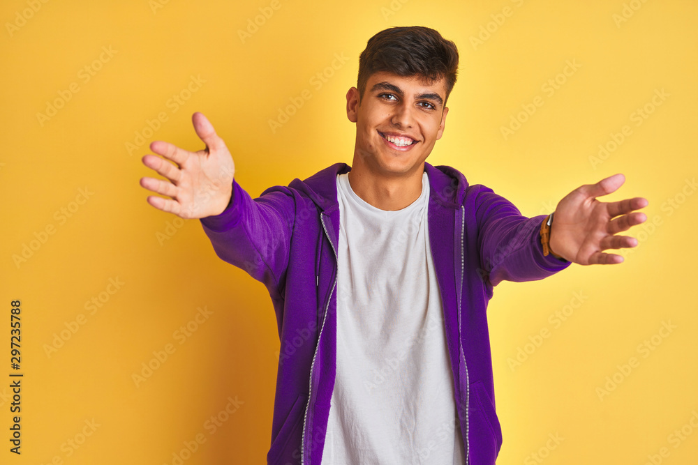 Young indian man wearing purple sweatshirt standing over isolated yellow background looking at the camera smiling with open arms for hug. Cheerful expression embracing happiness.