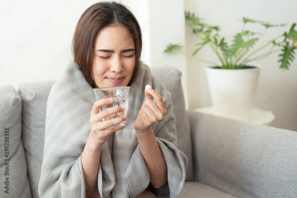 Asian Cute of  girl having headache and high temperature from illness holding pill of medicine and water sitting on sofa at home, She dislike to have a medicine, Health and illness concepts