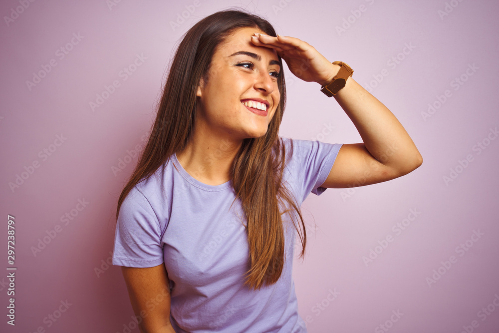 Young beautiful woman wearing casual t-shirt standing over isolated pink background very happy and smiling looking far away with hand over head. Searching concept.
