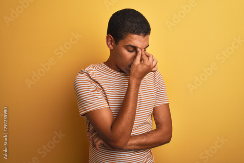Young handsome arab man wearing striped t-shirt standing over isolated yellow background tired rubbing nose and eyes feeling fatigue and headache. Stress and frustration concept. © Krakenimages.com