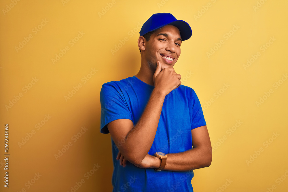 Young handsome arab delivery man standing over isolated yellow background looking confident at the camera smiling with crossed arms and hand raised on chin. Thinking positive.