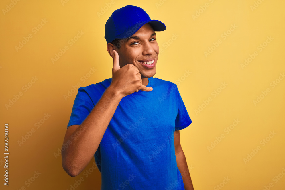 Young handsome arab delivery man standing over isolated yellow background smiling doing phone gesture with hand and fingers like talking on the telephone. Communicating concepts.