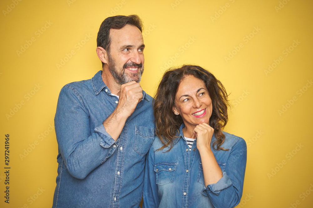Beautiful middle age couple together standing over isolated yellow background with hand on chin thinking about question, pensive expression. Smiling and thoughtful face. Doubt concept. Beautiful middle age couple together standing over isolated yellow background with hand on chin thinking about question, pensive expression. Smiling and thoughtful face. Doubt concept.