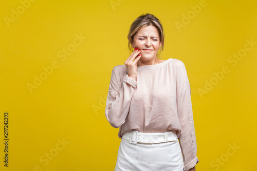 Portrait of upset sick young woman with fair hair in casual blouse standing with closed eyes, touching cheek suffering dental teeth ache, mouth injury. indoor studio shot isolated on yellow background © khosrork