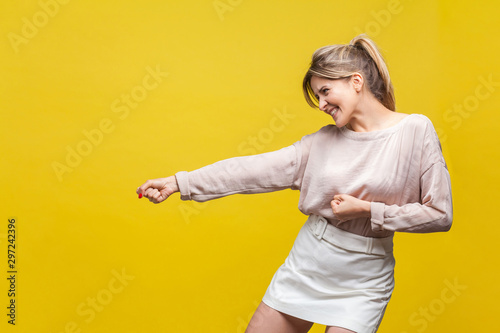 Profile side view of positive assertive young woman with fair hair in casual beige blouse standing with pulling gesture, much effort face expression. indoor studio shot isolated on yellow background