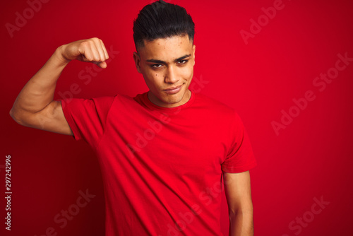 Young brazilian man wearing t-shirt standing over isolated red background Strong person showing arm muscle, confident and proud of power © Krakenimages.com