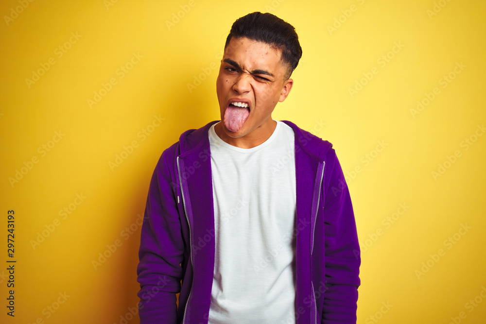 Young brazilian man wearing purple sweatshirt standing over isolated yellow background sticking tongue out happy with funny expression. Emotion concept. - obrazy, fototapety, plakaty 