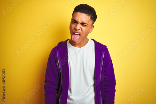Young brazilian man wearing purple sweatshirt standing over isolated yellow background sticking tongue out happy with funny expression. Emotion concept.