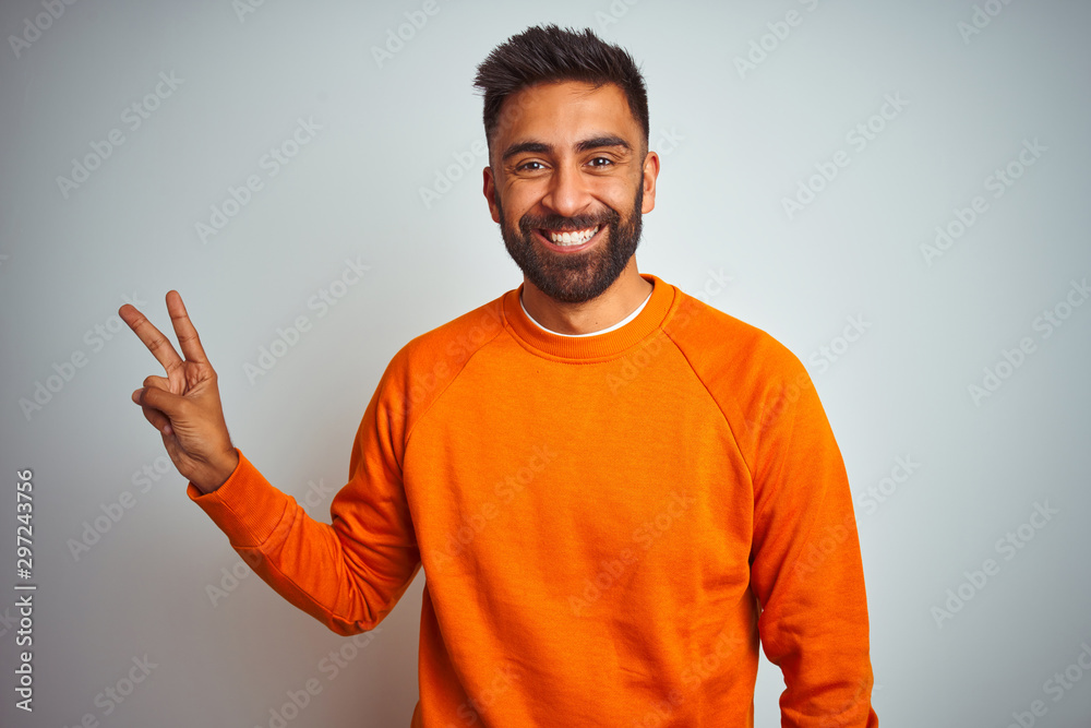 Young indian man wearing orange sweater over isolated white background smiling with happy face winking at the camera doing victory sign. Number two. Young indian man wearing orange sweater over isolated white background smiling with happy face winking at the camera doing victory sign. Number two.
