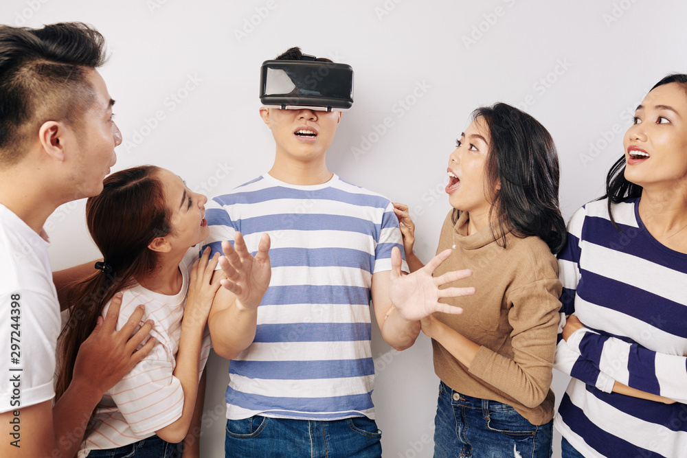 Shocked excited young Vietnamese people looking at their friend playing game in virtual reality glasses Shocked excited young Vietnamese people looking at their friend playing game in virtual reality glasses
