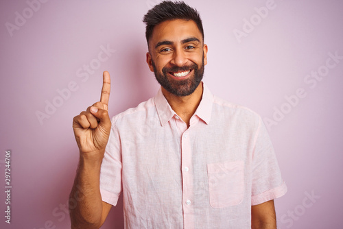 Young indian man wearing casual shirt standing over isolated pink background showing and pointing up with finger number one while smiling confident and happy.