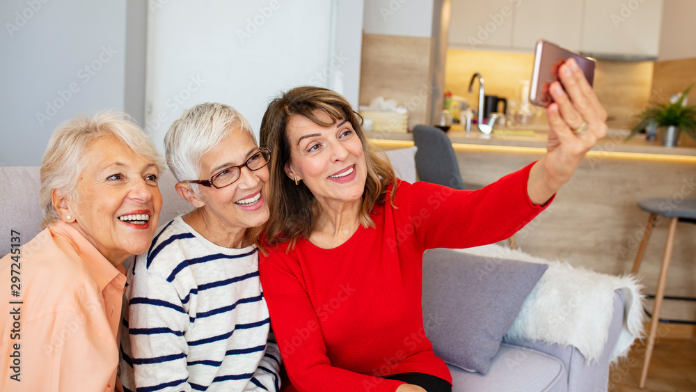 Obraz premium Beautiful senior women having fun taking selfies at home. Senior friends using a Phone. Senior women having fun with smartphone making selfies. Capturing their friendship