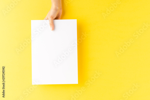 Young woman’s hand showing blank paper sheet on yellow desktop. Top view