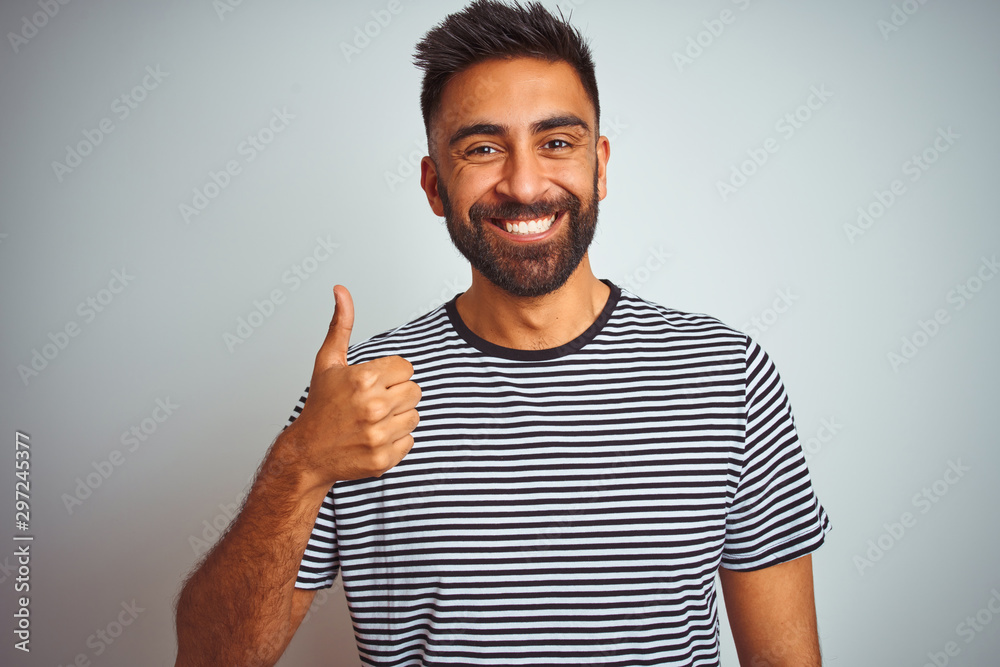 Young indian man wearing black striped t-shirt standing over isolated white background doing happy thumbs up gesture with hand. Approving expression looking at the camera with showing success. Young indian man wearing black striped t-shirt standing over isolated white background doing happy thumbs up gesture with hand. Approving expression looking at the camera with showing success.