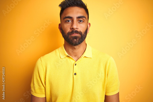Young indian man wearing polo standing over isolated yellow background with serious expression on face. Simple and natural looking at the camera.