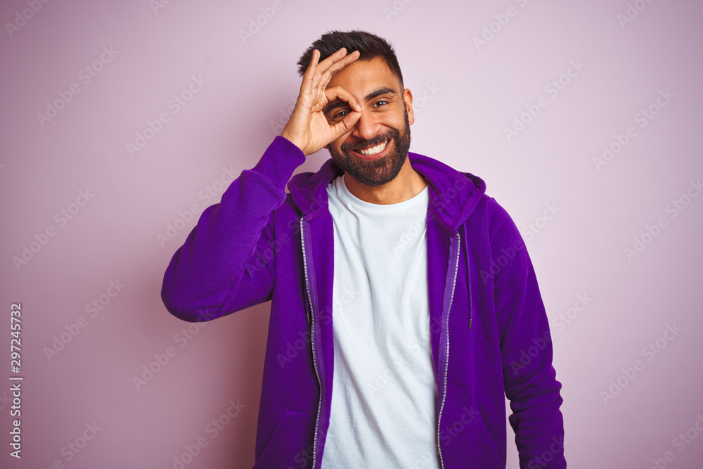 Young indian man wearing purple sweatshirt standing over isolated pink background doing ok gesture with hand smiling, eye looking through fingers with happy face.
