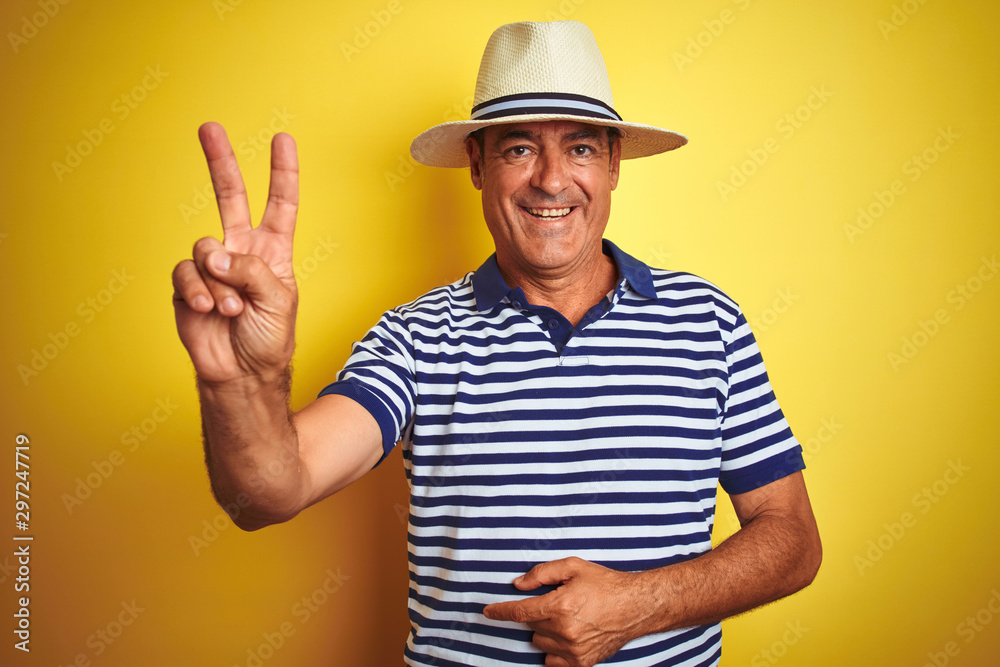 Handsome middle age man wearing striped polo and hat over isolated yellow background smiling with happy face winking at the camera doing victory sign. Number two.