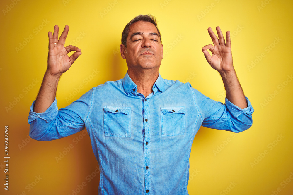 Handsome middle age man wearing denim shirt standing over isolated yellow background relax and smiling with eyes closed doing meditation gesture with fingers. Yoga concept.