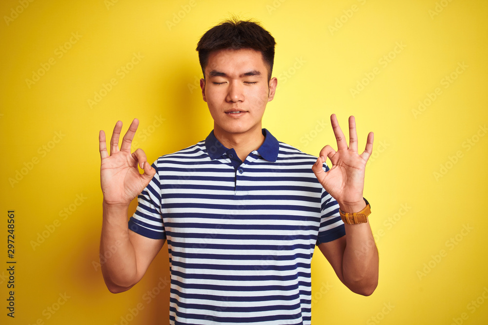 Young asian chinese man wearing striped polo standing over isolated yellow background relax and smiling with eyes closed doing meditation gesture with fingers. Yoga concept. Young asian chinese man wearing striped polo standing over isolated yellow background relax and smiling with eyes closed doing meditation gesture with fingers. Yoga concept.