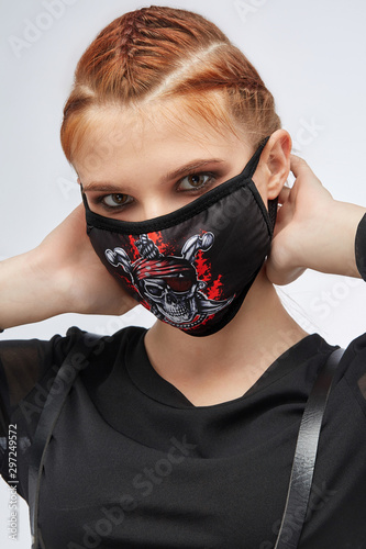 Close-up portrait of a ginger girl with three french braids, posing on a grey background. She's wearing black sweatshirt and leather belt. Her face is hidden by black mask with the pirate skull photo