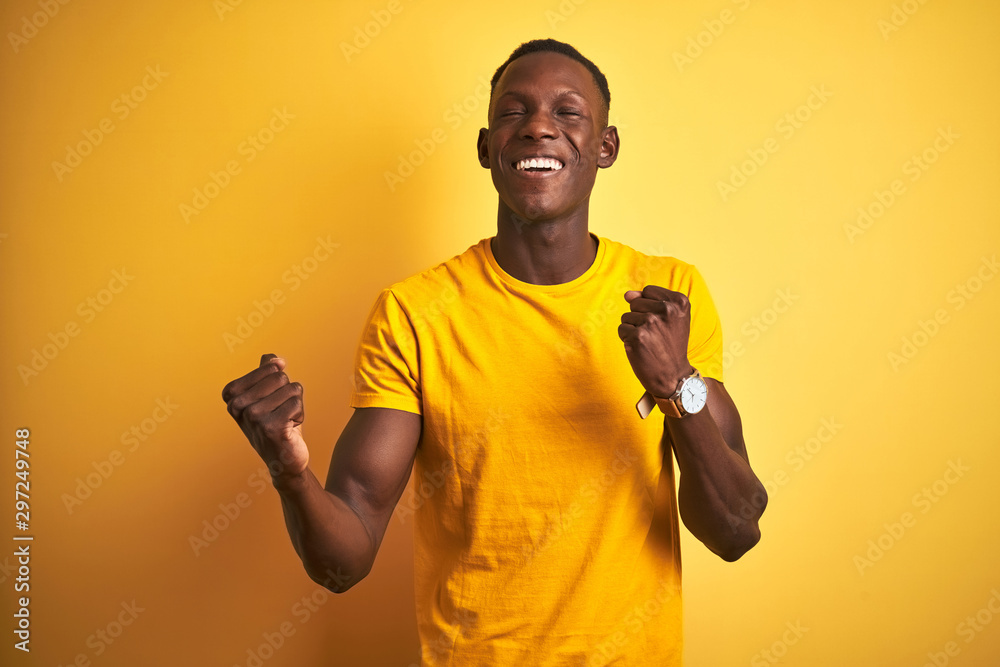 Young african american man wearing casual t-shirt standing over isolated yellow background celebrating surprised and amazed for success with arms raised and eyes closed. Winner concept. Young african american man wearing casual t-shirt standing over isolated yellow background celebrating surprised and amazed for success with arms raised and eyes closed. Winner concept.