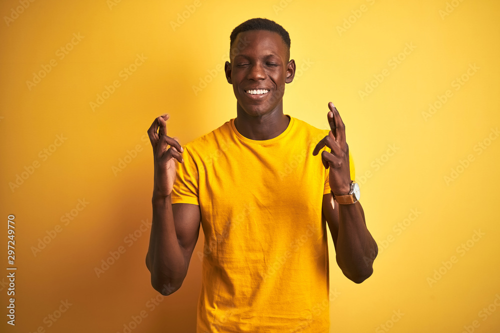 Young african american man wearing casual t-shirt standing over isolated yellow background gesturing finger crossed smiling with hope and eyes closed. Luck and superstitious concept. Young african american man wearing casual t-shirt standing over isolated yellow background gesturing finger crossed smiling with hope and eyes closed. Luck and superstitious concept.