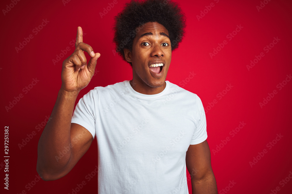 Young american man with afro hair wearing white t-shirt standing over isolated red background pointing finger up with successful idea. Exited and happy. Number one. Young american man with afro hair wearing white t-shirt standing over isolated red background pointing finger up with successful idea. Exited and happy. Number one.