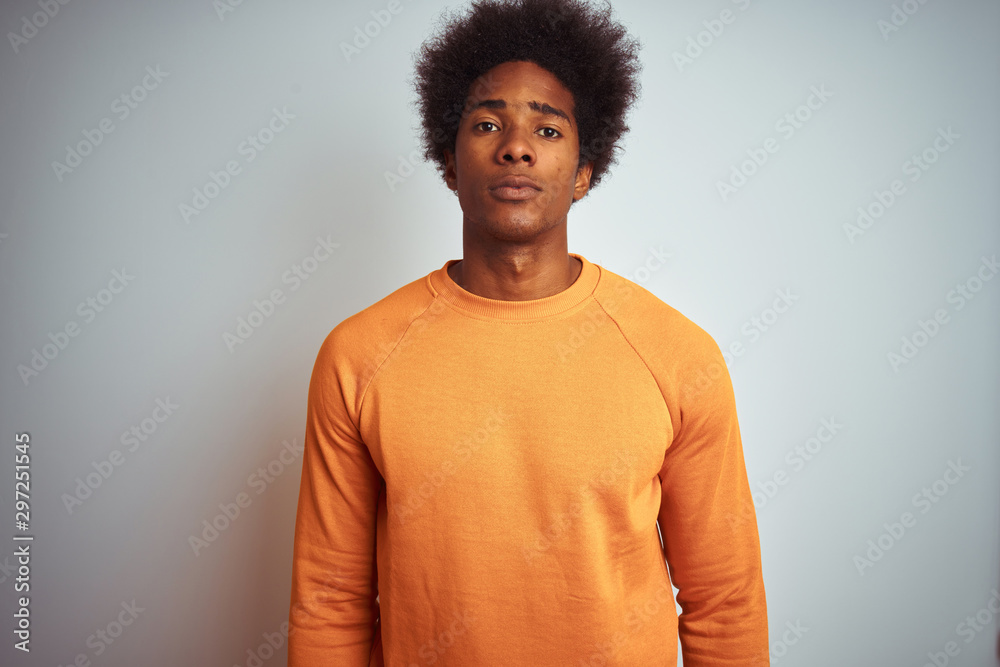 Young american man with afro hair wearing orange sweater over isolated white background with serious expression on face. Simple and natural looking at the camera. Young american man with afro hair wearing orange sweater over isolated white background with serious expression on face. Simple and natural looking at the camera.