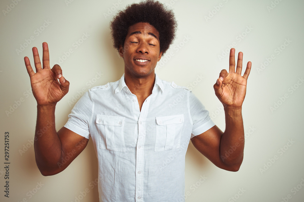 African american man with afro hair wearing shirt standing over isolated white background relax and smiling with eyes closed doing meditation gesture with fingers. Yoga concept.