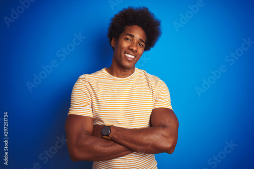 American man with afro hair wearing striped yellow t-shirt over isolated blue background happy face smiling with crossed arms looking at the camera. Positive person.