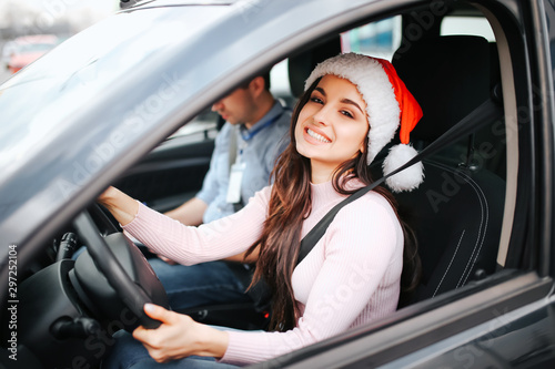 Picture of attractive young woman sit on driver's place. Hold hands on steering wheel and smile. Christmas or new year time. Wear red hat and smile. Man sit beside.