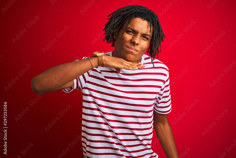 Young afro man with dreadlocks wearing striped t-shirt standing over isolated red background cutting throat with hand as knife, threaten aggression with furious violence Young afro man with dreadlocks wearing striped t-shirt standing over isolated red background cutting throat with hand as knife, threaten aggression with furious violence