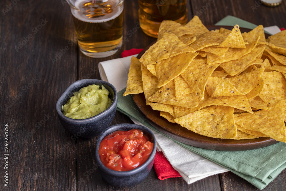 Famous Mexican snack, nachos with guacamole, red chilli sauce and beer on wooden table. Mexican cuisine background Famous Mexican snack, nachos with guacamole, red chilli sauce and beer on wooden table. Mexican cuisine background