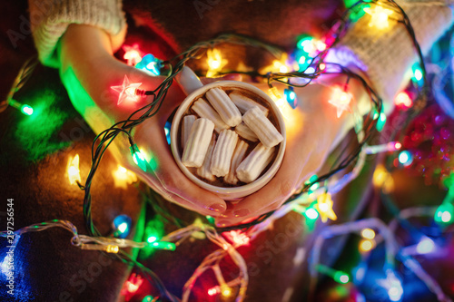 Fototapeta Naklejka Na Ścianę i Meble -  Girls hands hold cup of homemade hot chocolate with white marshmallows, a lot of lights of stars garland and blanket, christmas and new year concept