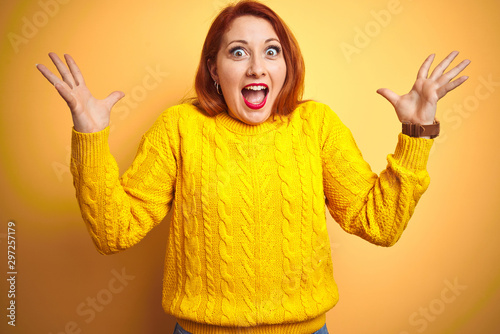 Beautiful redhead woman wearing winter sweater standing over isolated yellow background celebrating crazy and amazed for success with arms raised and open eyes screaming excited. Winner concept