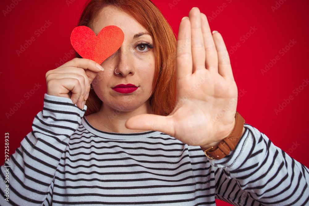 Young redhead romantic woman holding heart over red isolated background with open hand doing stop sign with serious and confident expression, defense gesture Young redhead romantic woman holding heart over red isolated background with open hand doing stop sign with serious and confident expression, defense gesture