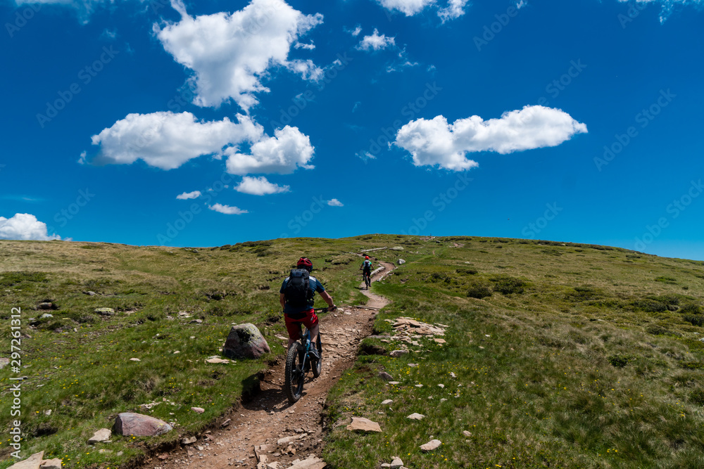 Zwei Mountainbiker auf einem Trail zwischen Wiesen aufwärts mit blauem Himmel und weißen Wolken