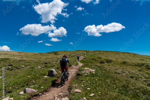 Zwei Mountainbiker auf einem Trail zwischen Wiesen aufwärts mit blauem Himmel und weißen Wolken