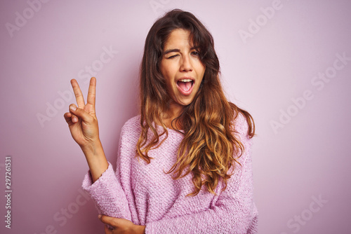 Young beautiful woman wearing sweater standing over pink isolated background smiling with happy face winking at the camera doing victory sign. Number two.