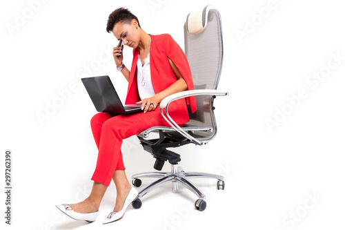 A young beautiful girl with short dark hair, makeup in a red office suit with bijuteria, expensive watches, sits in a computer chair with a laptop and a phone in hand photo
