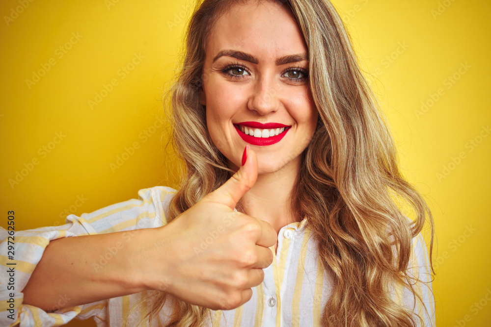 Young beautiful woman wearing stripes shirt standing over yellow isolated background happy with big smile doing ok sign, thumb up with fingers, excellent sign Young beautiful woman wearing stripes shirt standing over yellow isolated background happy with big smile doing ok sign, thumb up with fingers, excellent sign
