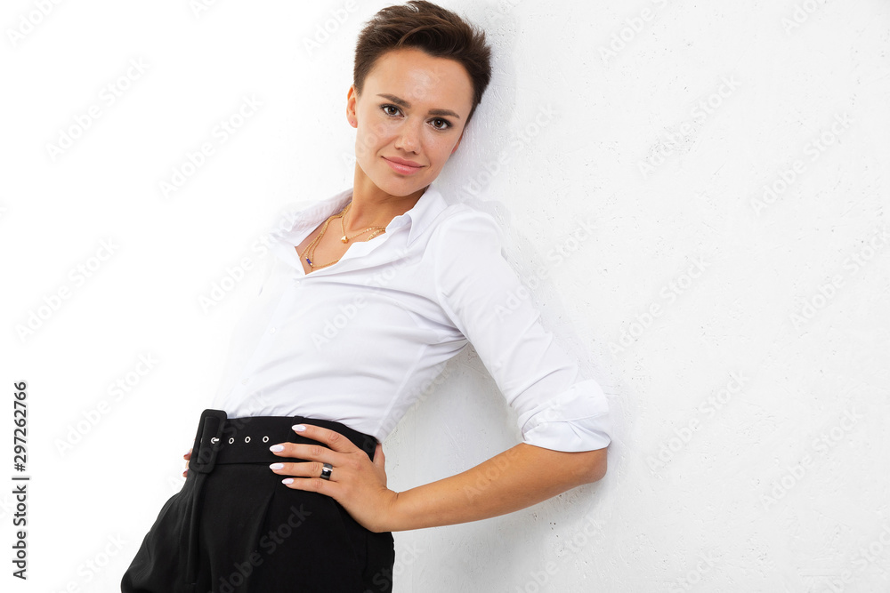 A young beautiful girl with short dark hair, makeup in a white shirt, black pants, with a wristwatch, bracelet and white manicure stands near a white wall and smiles A young beautiful girl with short dark hair, makeup in a white shirt, black pants, with a wristwatch, bracelet and white manicure stands near a white wall and smiles