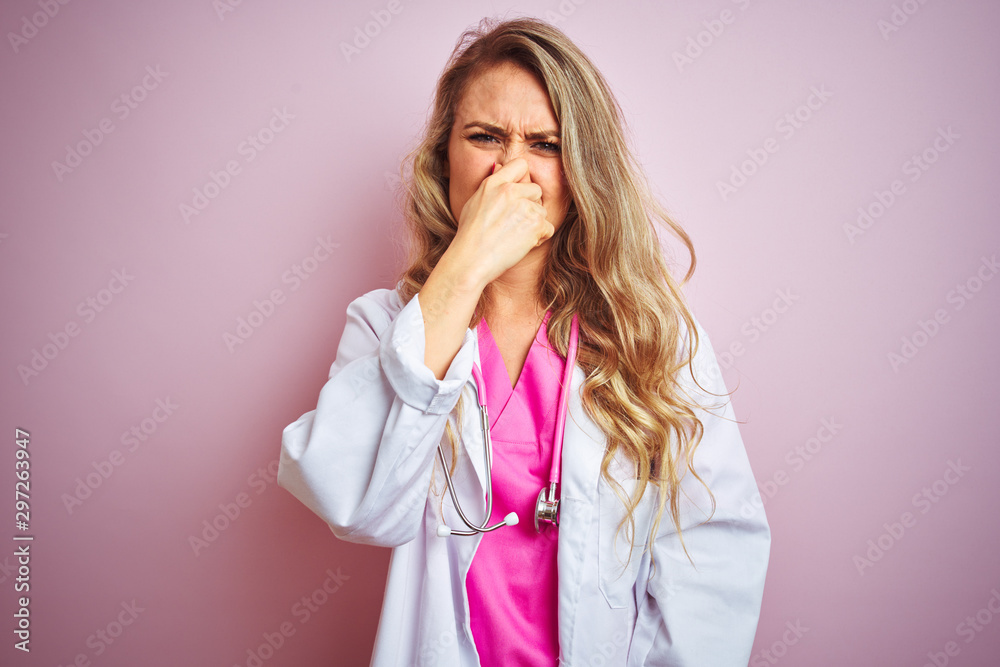 Young beautiful doctor woman using stethoscope over pink isolated background smelling something stinky and disgusting, intolerable smell, holding breath with fingers on nose. Bad smells concept. Young beautiful doctor woman using stethoscope over pink isolated background smelling something stinky and disgusting, intolerable smell, holding breath with fingers on nose. Bad smells concept.