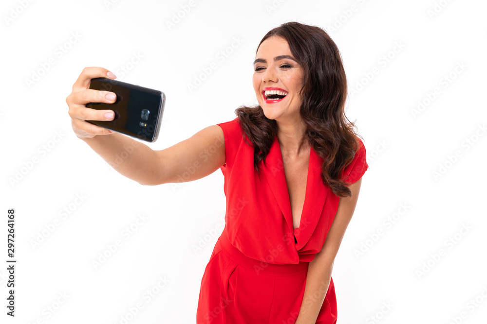 A young woman with red lips, bright makeup, a dazzling smile, dark wavy long hair, in a red summer dress stands and takes a selfie on her phone A young woman with red lips, bright makeup, a dazzling smile, dark wavy long hair, in a red summer dress stands and takes a selfie on her phone