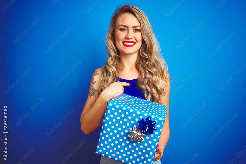 Young beautiful woman holding birthday present over blue isolated background with surprise face pointing finger to himself Young beautiful woman holding birthday present over blue isolated background with surprise face pointing finger to himself