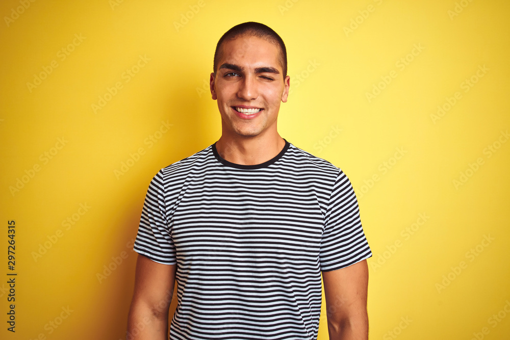 Young handsome man wearing striped t-shirt over yellow isolated background winking looking at the camera with sexy expression, cheerful and happy face. Young handsome man wearing striped t-shirt over yellow isolated background winking looking at the camera with sexy expression, cheerful and happy face.